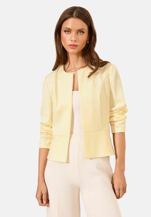 Woman with long brown hair wearing a pale yellow open-front jacket and light beige pants, standing against a plain background.