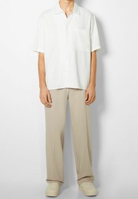 Camicetta bianca a maniche corte con chiusura a bottoni e tasca, abbinata a pantaloni beige a vestibilità rilassata e sneaker color crema chiaro.