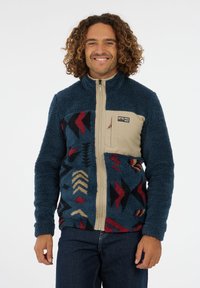Mann mit lockigem Haar, der eine blaue, gemusterte Fleecejacke mit beigem Brusttasche und Reißverschluss trägt und vor einem weißen Hintergrund lächelt.
