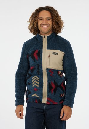 Man met krullend haar, gekleed in een blauwe fleecejack met patroon en een beige borstzak en rits, lachend tegen een witte achtergrond.