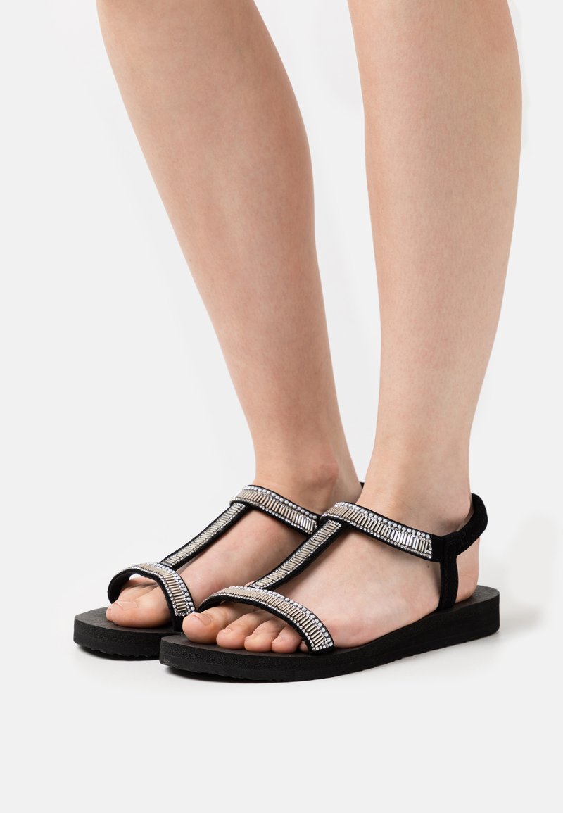 Svarte sandaler med T-stroppdesign, med en teksturert metallisk detalj og flat såle. Justerbar ankelstropp for passform og støtte.
