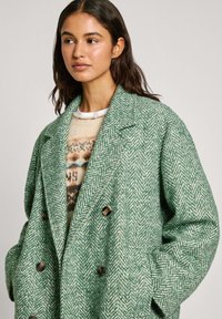 Manteau vert à motifs chevrons avec un large col, des boutons double boutonnage et un tissu texturé, porté par-dessus un pull crème à motifs.
