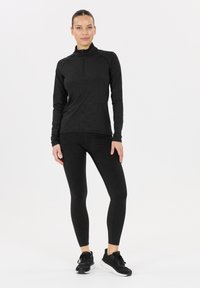 Vrouw staat in een zwart lange mouw, met rits, top en bijpassende leggings met zwarte sneakers tegen een effen witte achtergrond.
