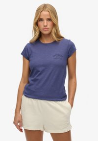 Mujer con cabello rubio, vistiendo una camiseta azul con logo y pantalones cortos casuales color crema, de pie con una mano en el bolsillo sobre un fondo blanco.