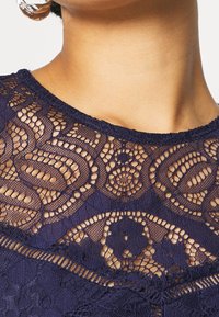 Námořnicky modrá krajka s propracovanými vzory a texturovaným designem. Výstřih je vroubkovaný a doplněný průsvitnou vrstvou.
