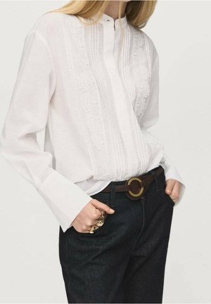 Kobieta nosząca białą bluzkę z długim rękawem z koronkowymi detalami, ciemne dżinsy, ciemnobrązowy pasek z okrągłą klamrą i kilka złotych pierścionków.