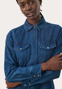 Dark blue denim shirt featuring a button-up design, long sleeves, two chest pockets, and silver hardware accents for a secure fit.