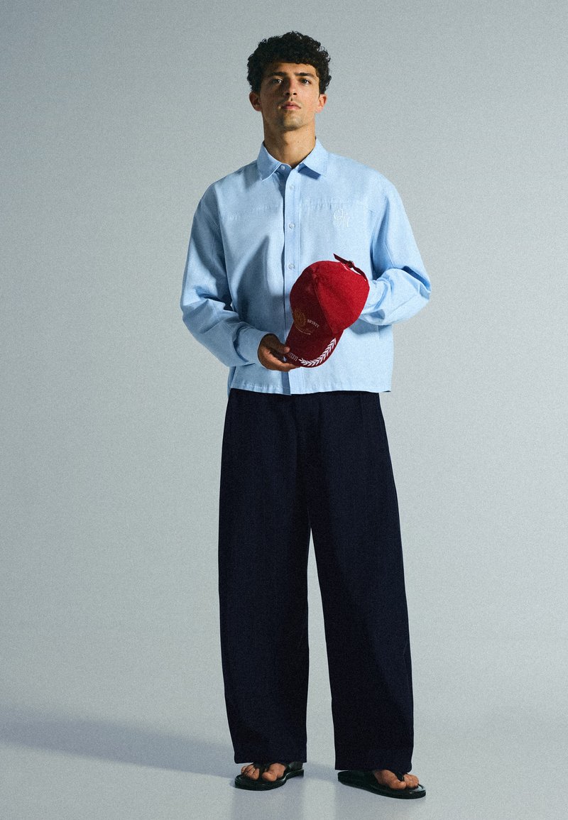 Jeune homme debout portant une chemise bleu clair, un pantalon large noir, des sandales noires, tenant une casquette de baseball rouge avec texte et design sur fond gris.