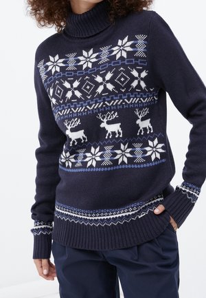 Pull-over à col roulé bleu marine avec des motifs de flocons de neige et de rennes blancs et bleus, porté avec un pantalon bleu marine assorti, main dans la poche.