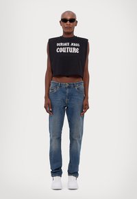 Fekete, cropped ujjatlan felső fehér "VERSACE JEANS COUTURE" felirattal, kék farmernadrággal és fehér tornacipővel párosítva. Sötét napszemüveg viselve.