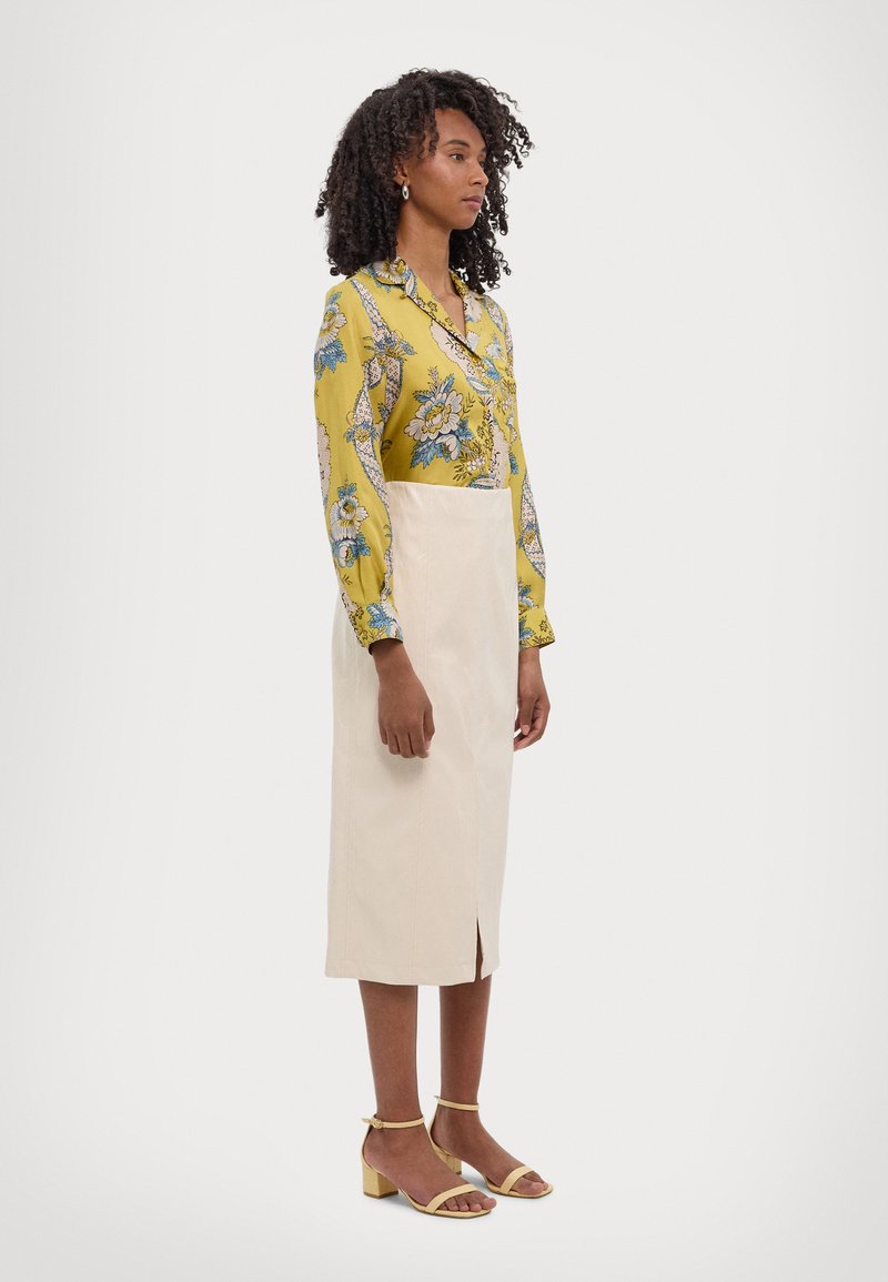 Gul blomstret bluse med lange ermer, kombinert med en lys beige skjørt med en frontslitt. Modellen bruker gule hælsandaler.