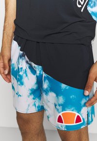 Černý sportovní top a vícebarevné plavkové šortky s modro-bílým vzorem mraků a detailem červeno-oranžového kulatého loga.