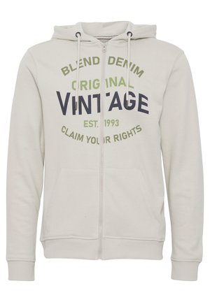 Vaaleanharmaa vetoketjuhuppari, joka on valmistettu pehmeästä kankaasta ja jossa on kengurun tasku. Etuosassa musta ja vihreä teksti: "BLEND DENIM ORIGINAL VINTAGE".