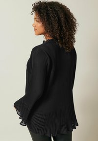 Schwarze gefaltete Bluse mit hohem Kragen, Rüschensaum und ausgestellten Ärmeln. Leichter Stoff mit glatter Textur und fließendem Design.