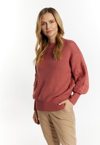 Pulover tricotat de culoare ruginită, cu tiv ondulat și mâneci texturate, pufoase. Asortat cu pantaloni bej deschis pentru un contrast atrăgător.