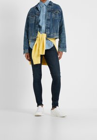 Giubbotto di jeans, camicia azzurro chiaro con bottoni, maglione giallo annodato in vita, jeans skinny scuri e sneakers bianche.