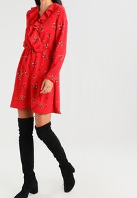 Abito rosso con colletto a volant e motivo floreale; indossato con stivali neri sopra il ginocchio. Tessuto morbido, design al ginocchio.