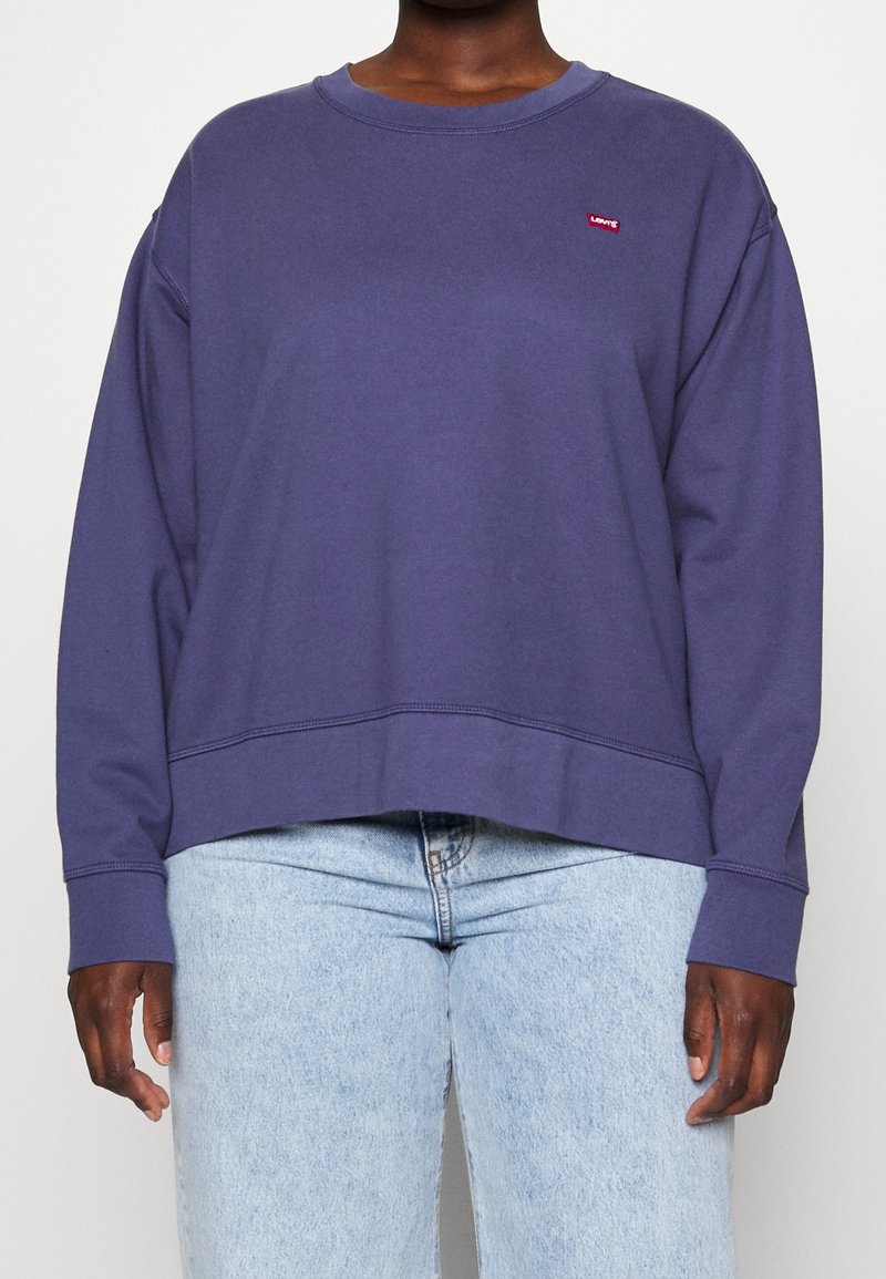 Personne portant un sweat-shirt violet uni à manches longues avec un petit logo rouge Levi's et un jean en denim bleu clair.