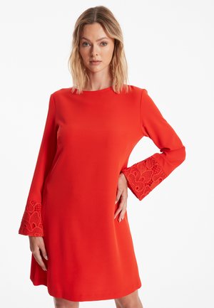 Robe rouge à manches longues avec des accents en dentelle aux poignets. Texture lisse, décolleté rond et coupe ample.