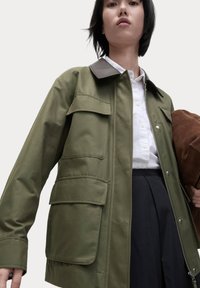Olijfgroene jas met een bruine kraag, meerdere zijzakken en een ritsluiting. Gestyled met een witte blouse en een zwarte rok.