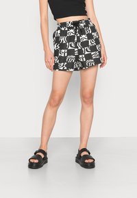 Femme portant un short à motif noir et blanc, un crop top noir et des sandales noires épaisses, debout sur un sol gris devant un mur blanc.