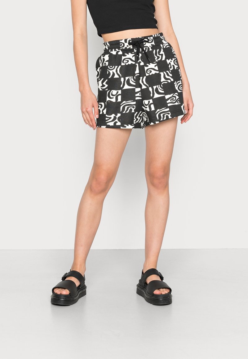 Femme portant un short à motif noir et blanc, un crop top noir et des sandales noires épaisses, debout sur un sol gris devant un mur blanc.
