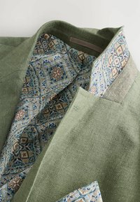 Grüne Blazer aus strukturiertem Stoff; das gefütterte Innenleben in Blau und Beige verfügt über geometrische Muster; enthält einen einfachen, klassischen Kragen und eine Tasche.