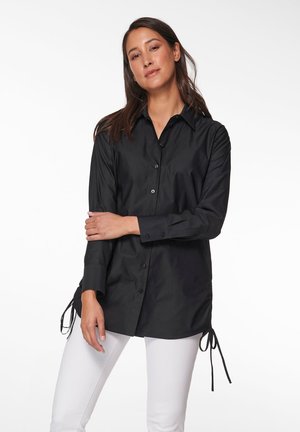 Zwarte blouse met lange mouwen, voorzien van zijkanten met strikken en een gladde textuur. Gecombineerd met witte broek voor contrast.