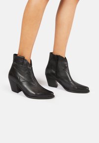 Bottes noires en cuir jusqu'à la cheville avec un bout pointu, une finition texturée et un talon bloc anglé. Dispose d'une fermeture éclair sur le côté et d'un motif de couture décoratif.