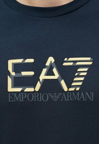 Tengerészkék pamut póló, texturált arany "EA7" logóval és az "EMPORIO ARMANI" felirattal, elegáns, modern betűtípussal.