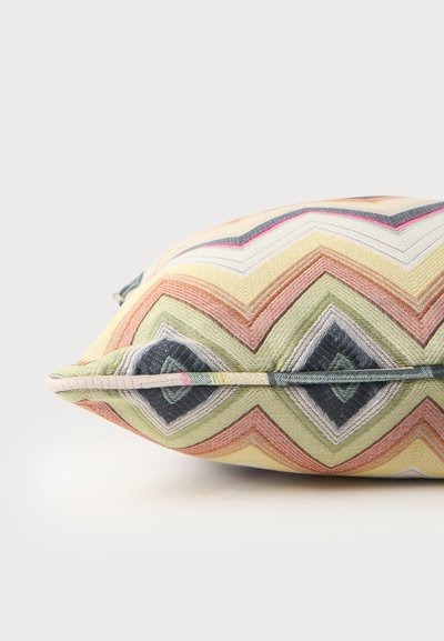 Coussin décoratif avec des motifs zigzag et losanges multicolores dans des tons pastel sur un fond clair.