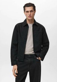 Črna denim jakna s ovratnikom, zadrgo in žepom na levi strani prsi, v kombinaciji s temnimi kavbojkami in svetlo sivo majico pod njo.