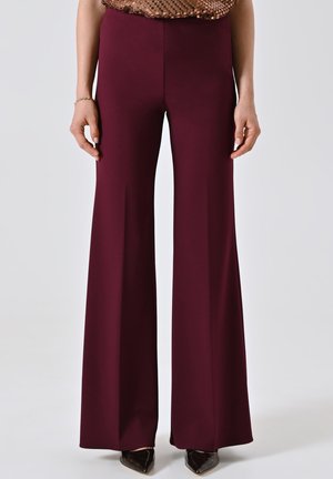 Donna che indossa pantaloni bordeaux a vita alta e gamba larga, abbinati a scarpe nere con punta e un top di paillettes bronzeo.