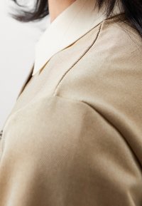 Světlý béžový blejzr s texturovanou tkaninou, který má kontrastní bílý límec, hladké švy a přiléhavý střih. Detailní záběr.