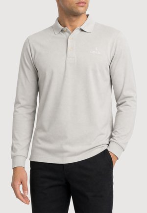Mann trägt hellgraues Langarm-Poloshirt mit drei Knöpfen und schwarze Hose, steht mit einer Hand in der Tasche.