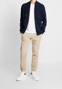 Marinblå strukturerad zip-jacka över en vit t-shirt, tillsammans med beige tapered joggerbyxor och vita sneakers med marinblå detaljer.