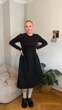 Czarna bluzka z długim rękawem zestawiona z czarną spódnicą z wysokim stanem, ozdobioną wiązaniami. Modelka ma na sobie czarne buty i białe skarpetki. Neutralne tło.