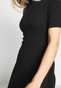 Robe noire à rayures, présentant une silhouette ajustée, des manches courtes et un tissu texturé. La robe a un col rond et une finition lisse.