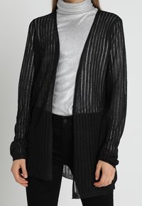 Černý kardigan s otevřeným předním dílem a vzorem svislých pruhů, lehká pletená textura, dlouhé rukávy a vrstvený design přes stříbrný rolák.