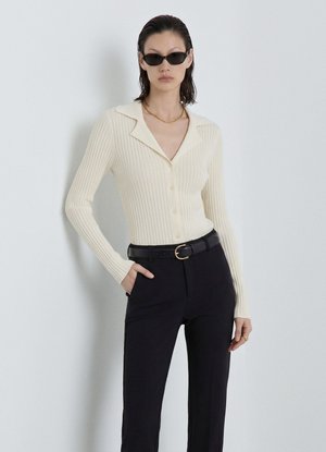 Frau trägt cremefarbenen, gerippten, geknöpften Cardigan, schwarze Hose mit Gürtel, Sonnenbrille und goldene Halskette, steht vor einer weißen Wand.