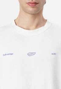 Weißes Baumwoll-T-Shirt mit lila gesticktem Logo und Text. Einfacher runder Ausschnitt, lockere Passform und glatte Textur.