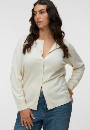 Kvinde med mørkt, bølget hår iført en off-white cardigan med knapper og blå jeans, stående med den ene hånd på hoften mod en ensfarvet baggrund.