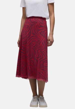 Donna che indossa una gonna midi a righe zebrate rosse e viola scuro, una maglietta bianca a maniche corte e sneakers bianche, in piedi su uno sfondo semplice.