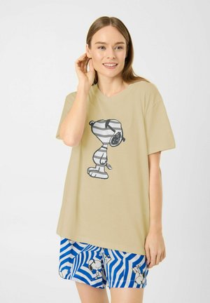 Femme souriante avec un T-shirt beige featuring un graphique rayé de Snoopy et un short bleu et blanc à motifs zigzags avec des impressions de Snoopy.