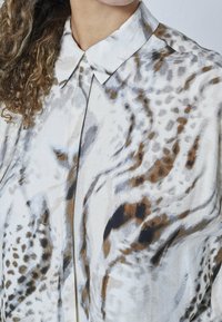 Letvægtig, kravebluse i creme, med et flydende mønster af brune og grå pletter. Stoffet har en blød, tekstureret overflade.