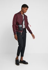 Burgunderröd bomberjacka med dragkedjedetaljer, grå t-shirt, svart axelremsväska, smal passform svarta byxor och svarta loafers med tofsar.