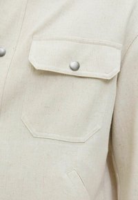 Kreminės spalvos striukė su tekstūruotu audiniu, turinti du kišenėlių užklotus ir sidabrinius spaudėjus. Išsiskiria paprastu ir funkcionaliu dizainu.
