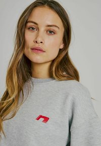 Grå sweatshirt med texturerad yta, med en röd logga på bröstet. Halsringningen och ärmsluten är ribbade för extra detalj.