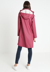 Bordowy płaszcz przeciwdeszczowy z kapturem, z białą podszewką. Długość do połowy uda, prosty krój i długie rękawy. Noszony z ciemnymi dżinsami i białymi trampkami.