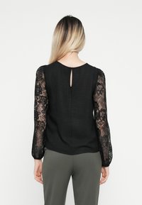 Svart topp med teksturert stoff og gjennomsiktige blonde lange ermer. Har en nøkkelhulldetalj bak og en rett snitt. Kombinert med khaki bukser.